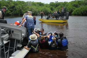 The crew of people holding the dolphin have placed it into a tarp for transport. They are lifting it up at the edge of an airboat that is tilted down towards them slightly. More people stand at the edge of the airboat to pull the dolphin up. The airboat that originally spotted the dolphin floats nearby.