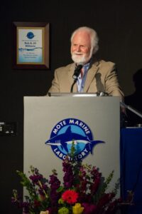 Dr. Michael P. Crosby, Mote President and CEO stands at a podium sporting a large Mote Logo. He smiles as he gives an address commemorating the life and legacy of Mote Founder Dr. Eugenie Clark. A beautiful bouquet of fresh flowers stands at its base, reaching as high as the table top.