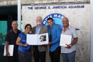 Genie's children and grandson pose with Mote President and CEO Dr. Michael Crosby by the shark tanks at Mote Aquarium. From left to right: Niki Konstantinou, Aya Konstantinou, Dr. Crosby, center, holding a picture of Dr. Eugenie Clark, in full dive gear despite her advanced age, Eli Weiss, and Tak Konstantinou. 