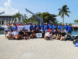 A group photo of at least 60 people gather at the dock with scuba gear and banners, getting ready to set out on their mission to restore the reef. Participants are a mix of Combat Wounded Veteran Challenge, SCUBAnauts International and Mote Staff members.