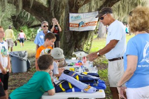 Kids lean on a table covered with gear related to ethical angling, including a mounted Snook and life jackets. An older man in sun glasses and a baseball cap talks to the kids about the items while other people look on. They are outside, under a large oak tree draped with Spanish Moss. 