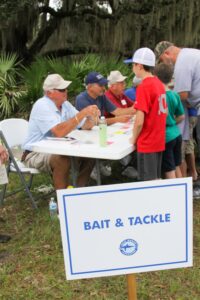 Several boys and a Dad stand at a table containing information cards talking with several old men in baseball caps seated on the other side. They are outside in the grass next to a sign proclaiming the table the "Bait and Tackle" station.