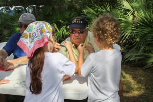 A young girl and and boy hold rubbery shrimp shaped fishing lures, examining them closely. An older man in a baseball cap and sunglasses assists them while another man assists another child out of frame. The booth is outside, surrounded by palmettos. 