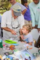 A little boy with curly blond hair watches intently as a Mote volunteer demonstrates how to tie a fly. The booth is strewn with fly parts and tools.