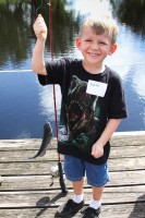 A small boy in a T-rex t-shirt proudly holds up the small catfish he caught, still hanging from the line.
