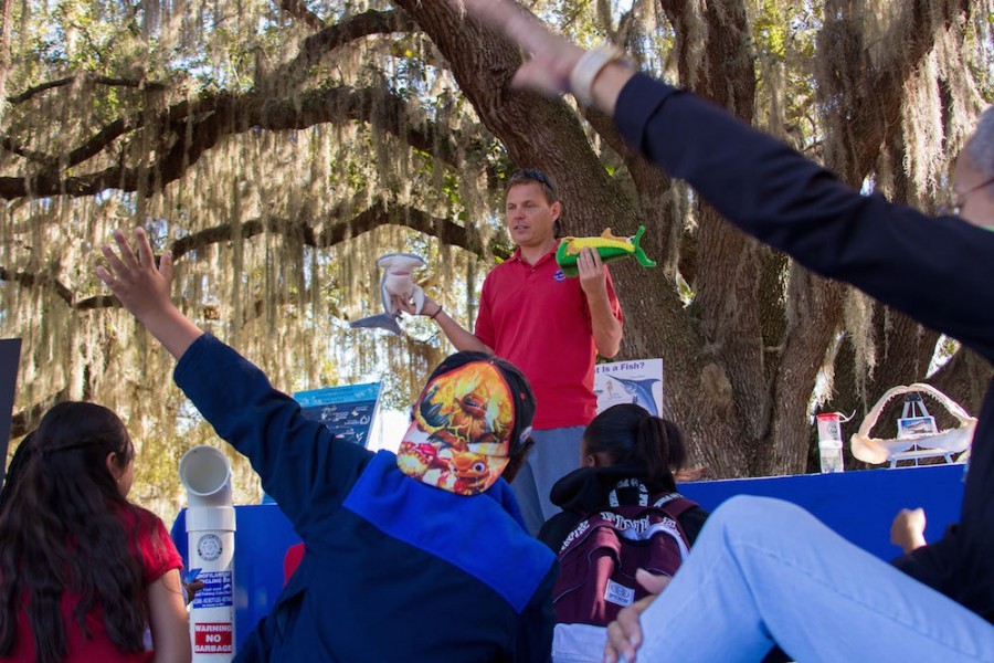 Mote staff member stands under a large oak tree, it's branches draped with Spanish moss. He poses a question to a group of elementary school students seated around him, while holding a toy stuffed shark in his right hand and a toy stuffed fish in his right for emphasis. A boy in a backwards baseball cap raises his hand.