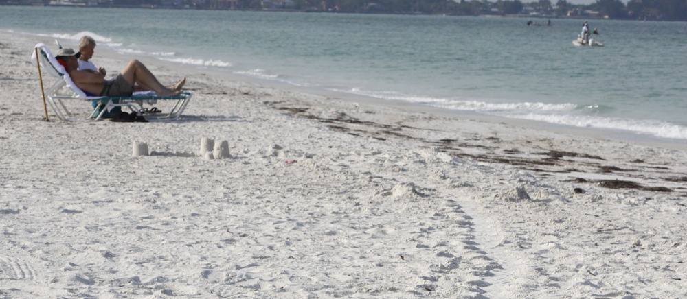 Panoramic of a typical Florida day at the beach, clear except for a small sand castle and small patches of seaweed. The surf is calm, two men rest in lawn chairs while a zodiac and kayaker fish offshore.