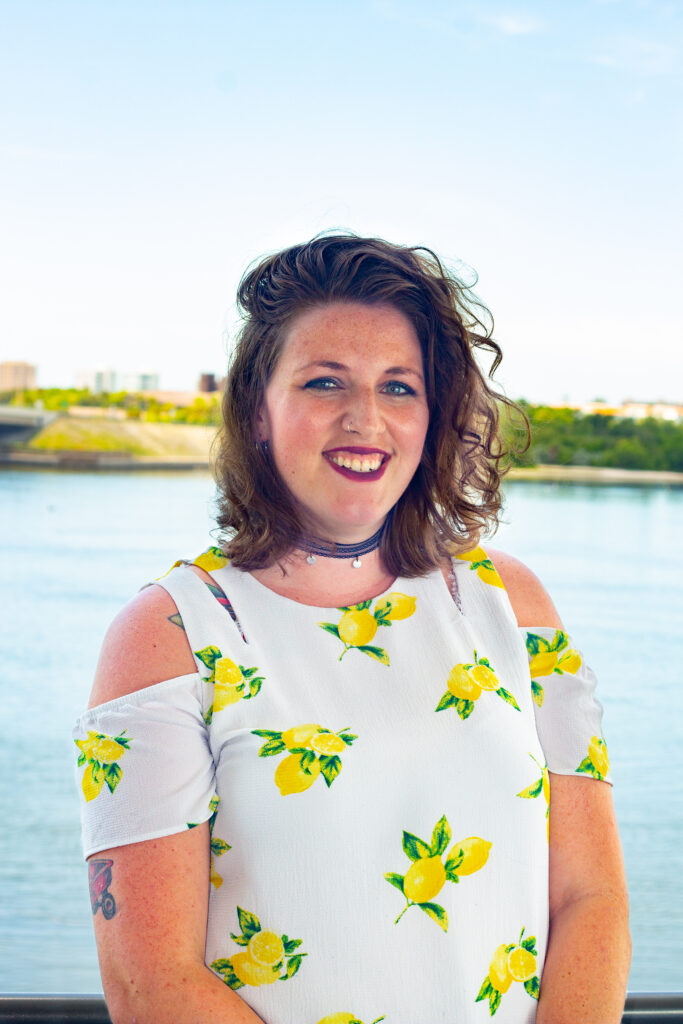 A portrait of Stephannie Kettle. She smiles at the camera in a bright floral patterned evening dress. Behind her is the bay flanked by the Sarasota skyline.