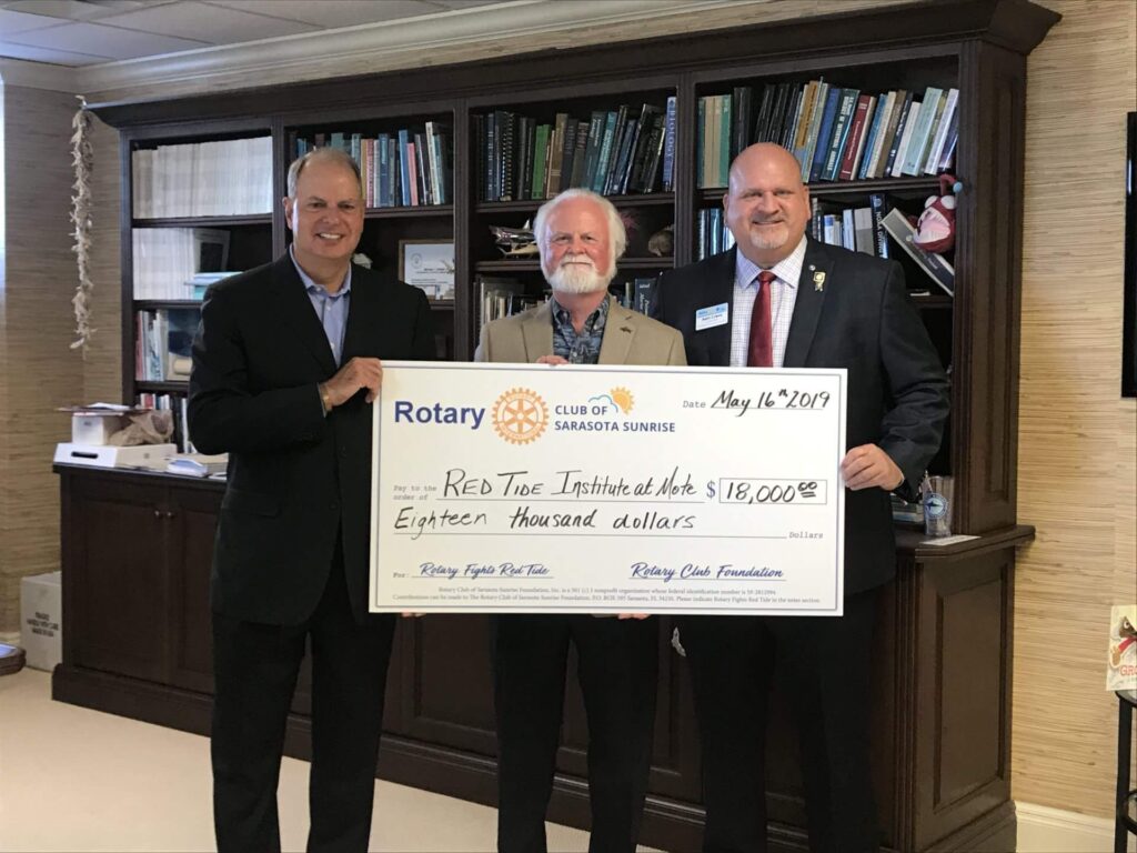 Three menin suites hold a large $18,000 check to the Red Tide Institute, curtesy of the Rotary Club of Sarasota Sunrise. The men are: Brian Maddox of Stifel, Mote Director Dr. Michael Crosby, and Andy Lyman of the Rotary Club of Sarasota Sunrise. They stand in front of a large wooden bookshelf at Mote, filled with scientific books, journals, models of aquatic life and a stuffed toy angler fish. ,