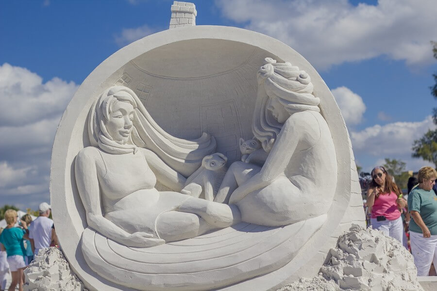 Sand sculpture depicting two women in dresses, sitting together and holding their pet cats.