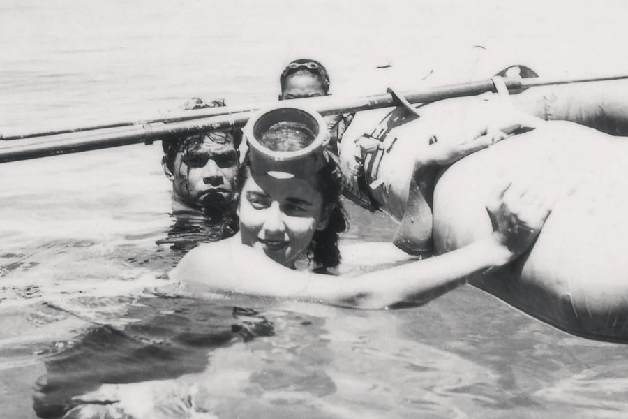 A young Eugenie Clark treds water next to a zodiac with oars. She has a circle dive mask pushed up on her forehead and is looking at something off to the side with a small smile. Two men tred water behind her. They all hold onto the side of the zodiac for stability. It is an old picture, all in black and white.