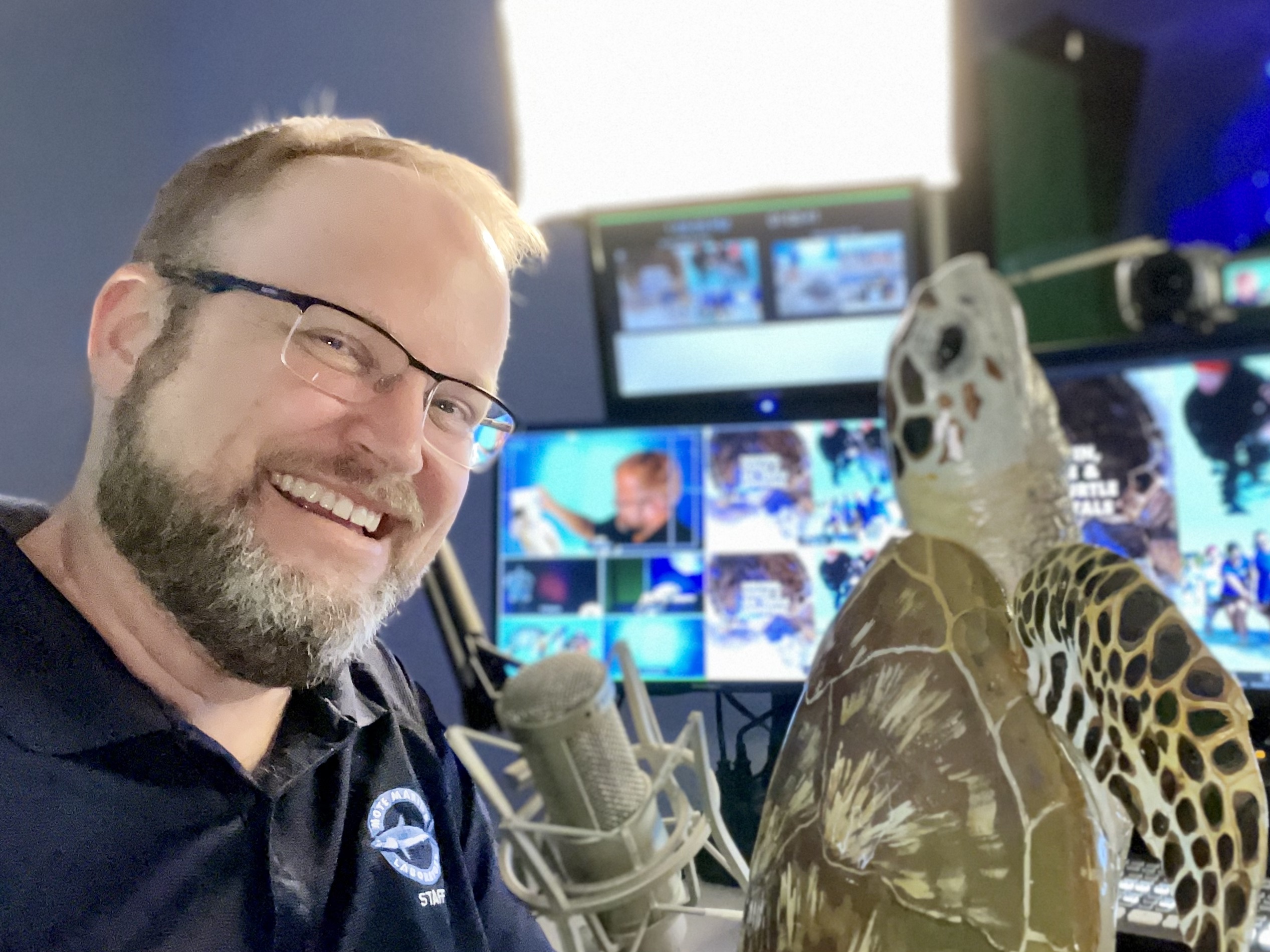 A smiling man with glasses and a beard, wearing a dark blue shirt with a logo, poses next to a model of a sea turtle in a studio. Behind him, there are computer monitors displaying various images and video feeds, with a bright light shining in the background.