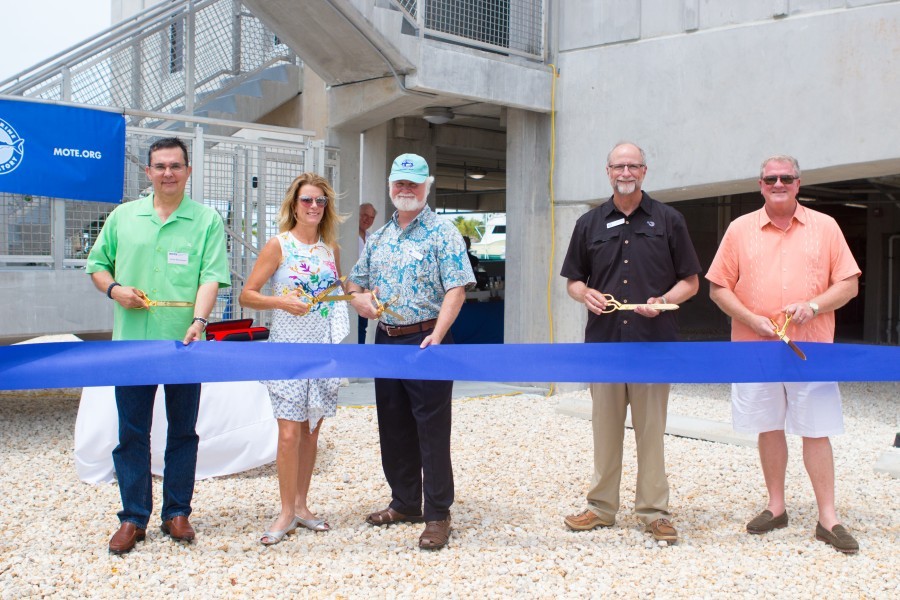 Five people stand behind a large blue ribbon that stretches across the entire picture. The middle person, a 60ish man with a white beard and blue patterned Hawaiian shirt and baseball cap is getting ready to cut it. Behind them is the newly constructed research center.