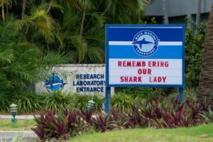 A large sign with the Mote Logo and the words "Remembering our Shark Lady" stands in front of the Mote Research Laboratory Entrance. The grounds are nicely decorated, full of palms, hibiscus, ornamental grasses and bromeliads.