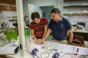 Intern Mauricio Rodrigues and Mote Scientist Dr. Richard Pierce work in the Ecotoxicoloy Lab together, reviewing something on a sheet of paper. The laboratory table they are working on is strewn with scientific equipment, including beakers of all sizes, tubing, air pumps, notebooks and clip boards. On the table to their right are several large glass jugs full of algae. Behind them is a fume hood, a large pressurized tank and a shelf covered in large sea shells that sits above a workstation. 