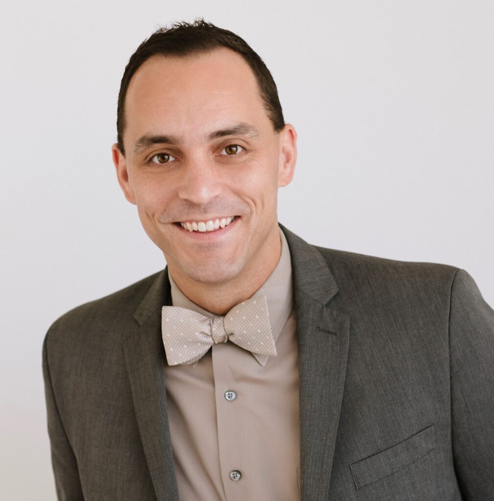 A portrait of new VP Kevin Cooper, smiling at the camera with close cropped hair wearing a grey suit and bow tie.