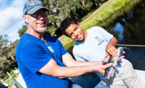 Dr. Brennan in a baseball cap and a 5th grade boy from Abel Elementary hold a foot long catfish the student caught at Mote's 10th annual Teach-A-Kid Fishing & Ecology Clinic.
