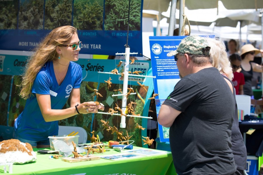 Staff Biologist Shelby Luce stands befind her booth covered with model staghorn corals. She holds one pecie as she speaks to a visiting couple. A replica of the antenna-like piping structure Mote uses to farm coral fragments in the sea hangs nearby.