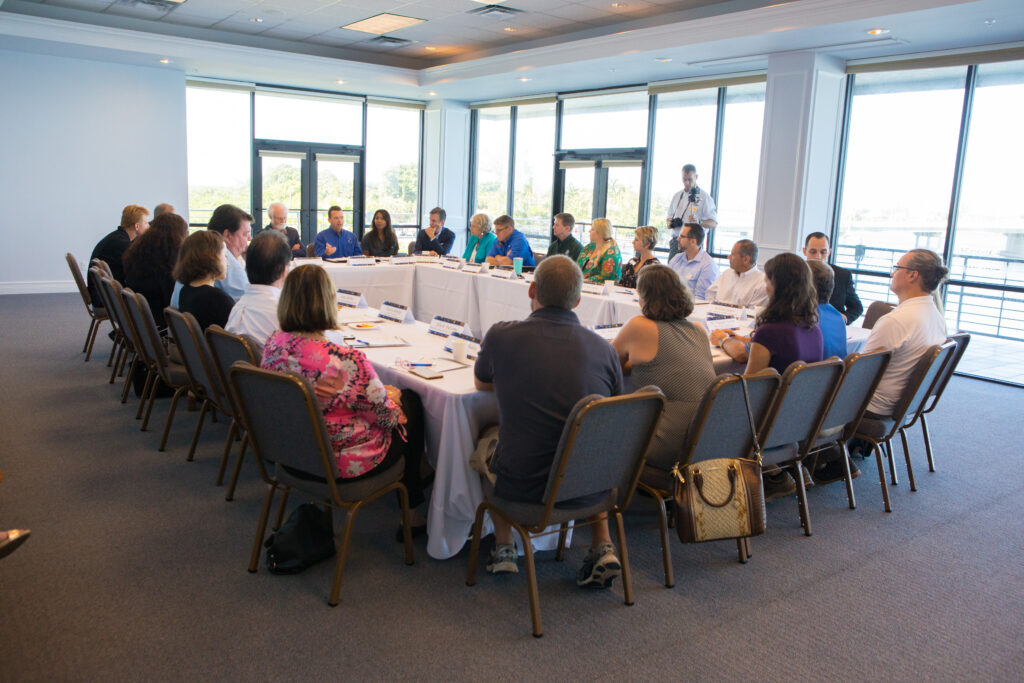 Leadership from mote and several state agencies meet to discuss a new partnership to address red tide. They sit at for long tables arranged in a square in a large glass room.