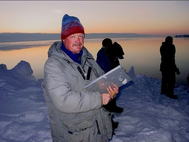 John Reynolds standing on an iceberg with several other scientists at dawn, in a grey parka and woolen hat, his face red with cold. He holds a metal clipboard and pencil.