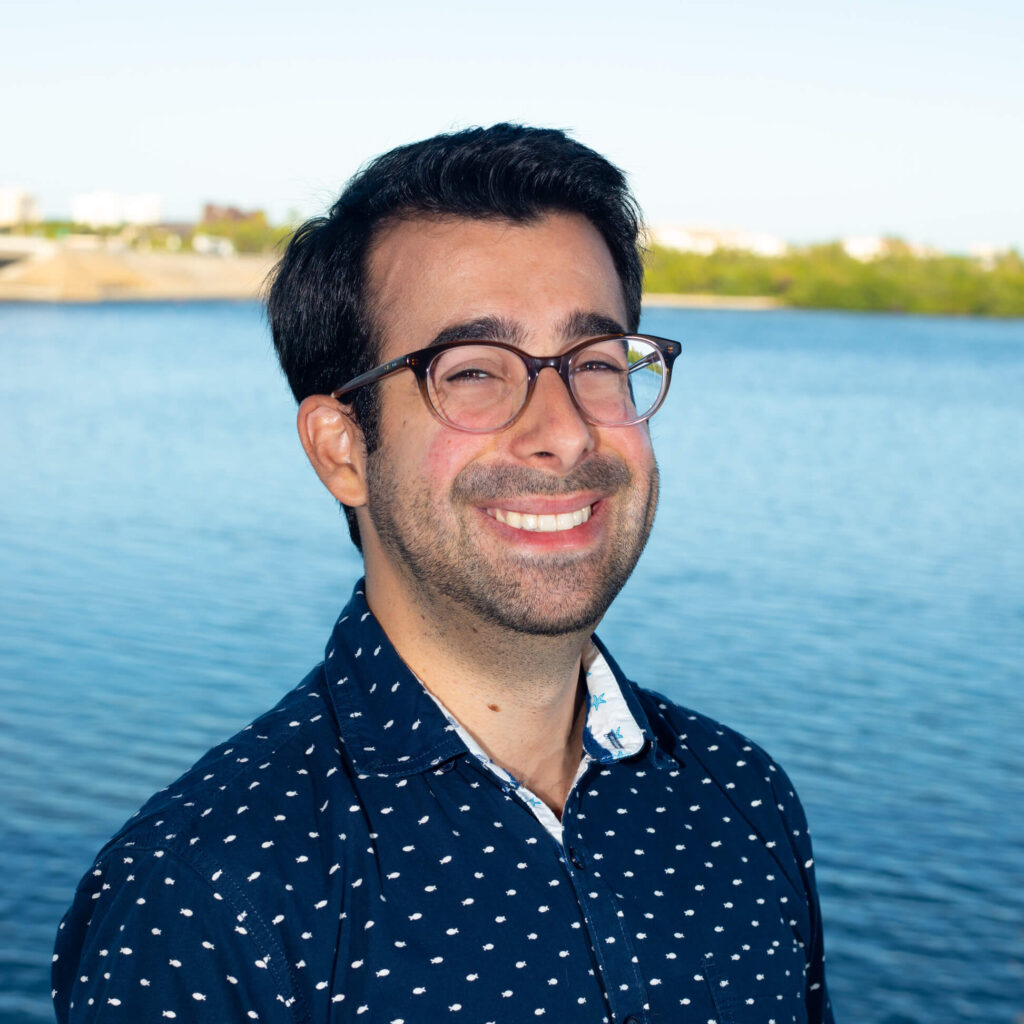 Portrait of Ross Johnston in front of Sarasota Bay. He has a broad smile, large glasses and short dark hair. His shirt is decorated with tiny fish that look like polka-dots until one looks closely.