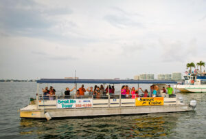 Guests wave from aboard the Sarasota Bay Explorers Eco-Tour Boat on Sarasota Bay. Signs on the boat highlight SeaLife Tours and Sunset Cruises.