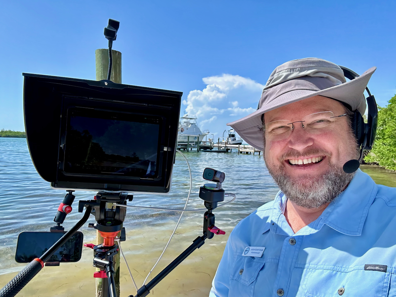 A man with glasses, a beard, and a wide-brimmed sun hat is smiling at the camera while wearing a headset microphone. He is standing on the shore of a body of water with clear blue skies and docks in the background. Next to him is a large camera setup on a tripod, which includes a teleprompter, a mounted microphone, and a connected smartphone. Mangrove trees are visible on the right side of the frame. The man appears to be broadcasting or recording a presentation near the water.