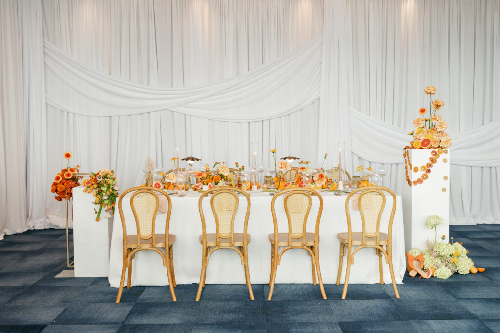 A long wedding table is set for a feast with orange florals set on a bright blue carpet and white draping behind it.