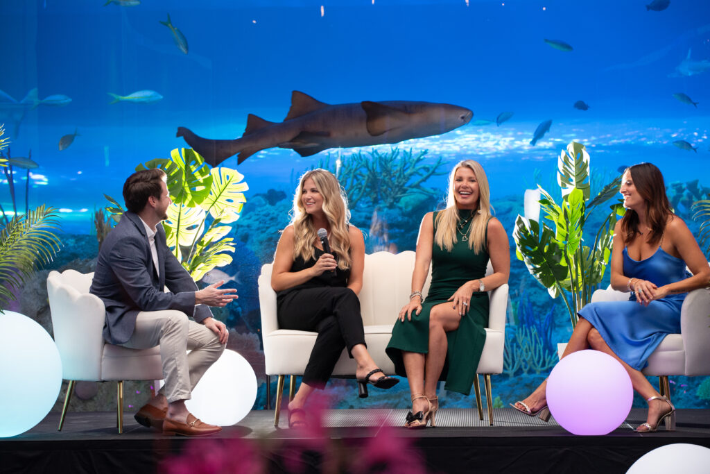 A panel sits ona stage in front of a large figh tank with a shark swimming above them