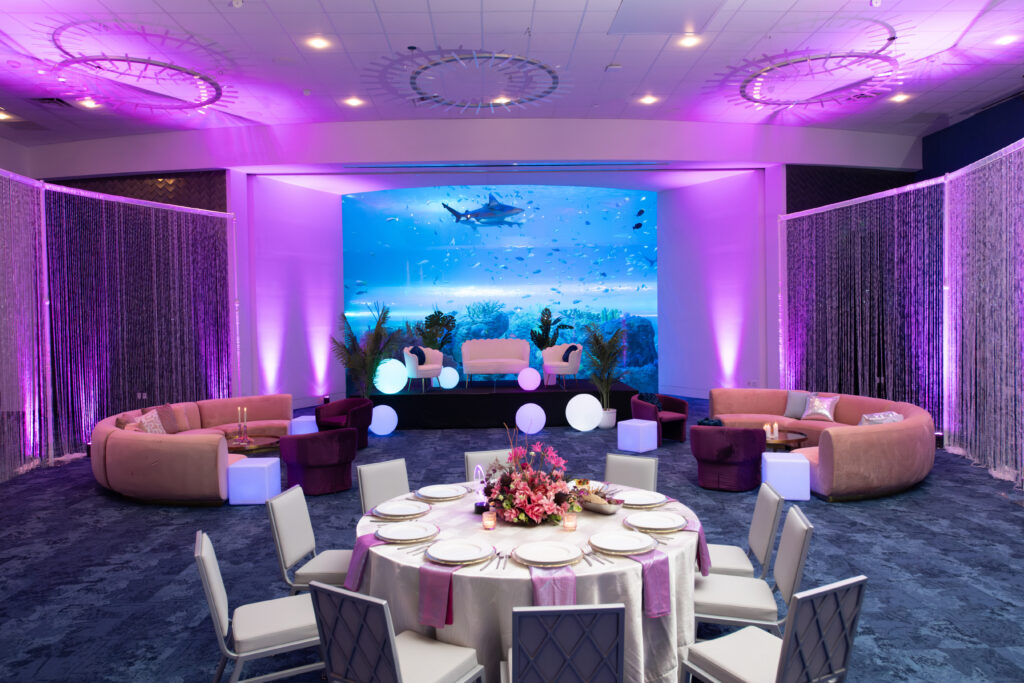 A large room features a floor to ceiling fish & shark tank, a stage, couches, and a banquet table set up with funky decor